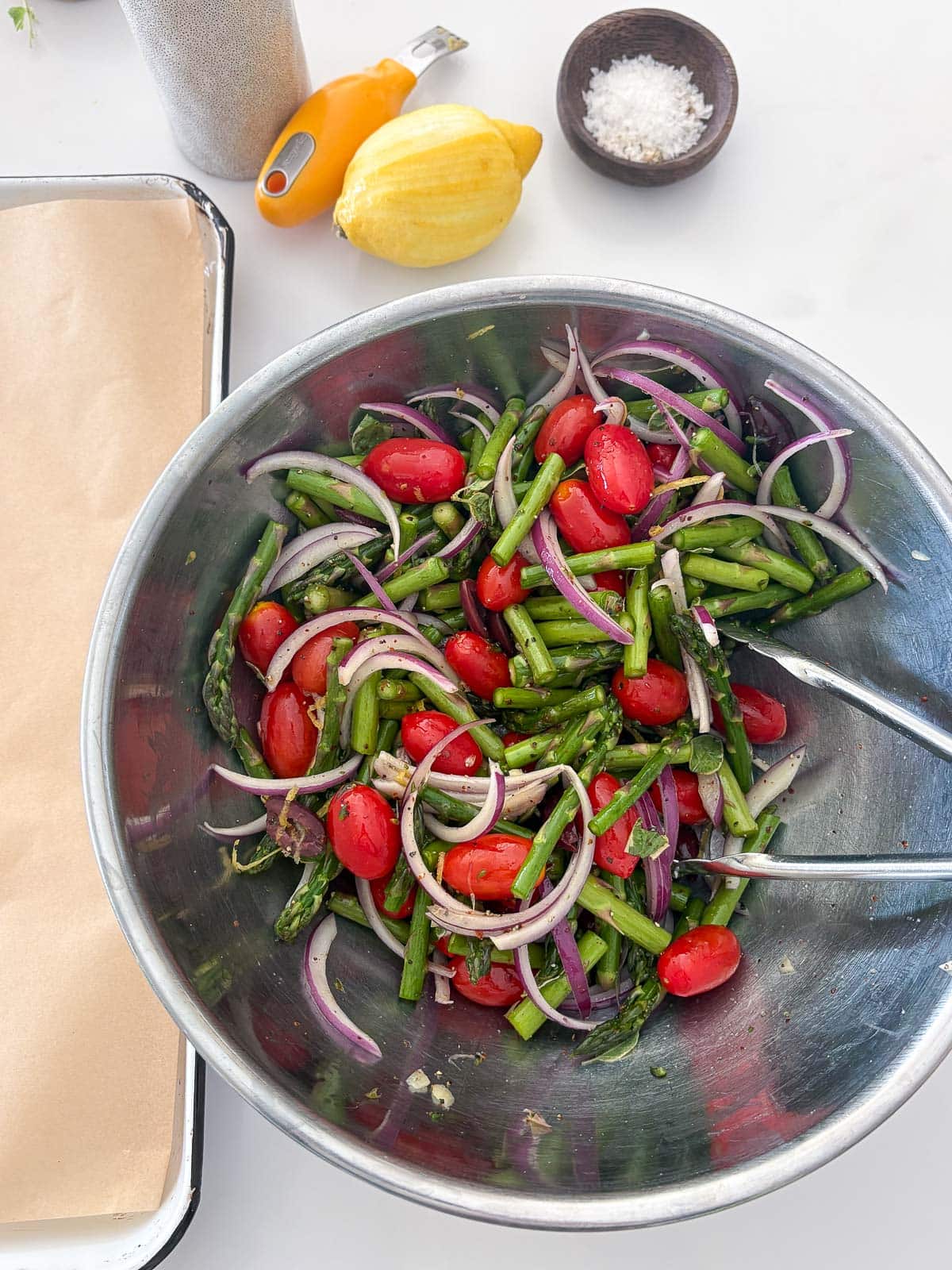 mixing bowl with tongs tossing asparagus, red onion, and cherry tomatoes. 