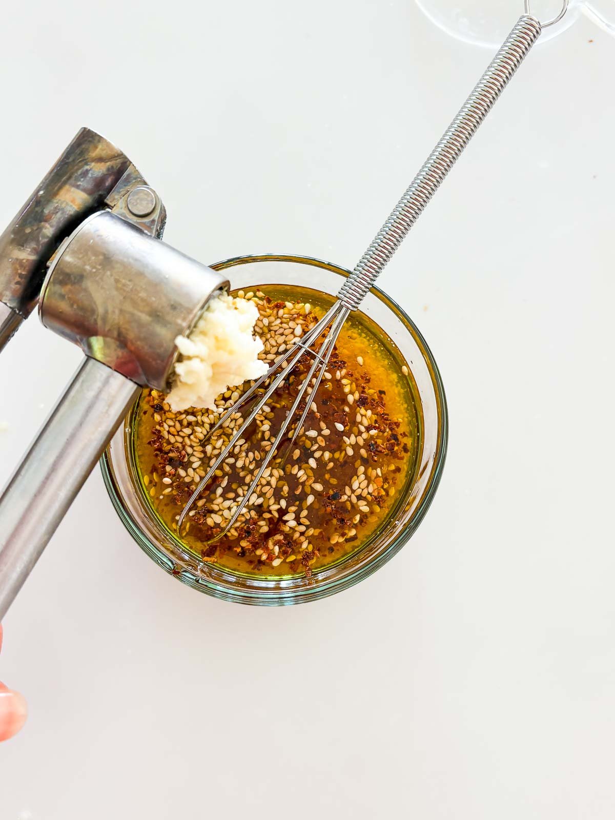 garlic press is pressing minced garlic into a jar of sesame ginger dressing with a small whisk. 