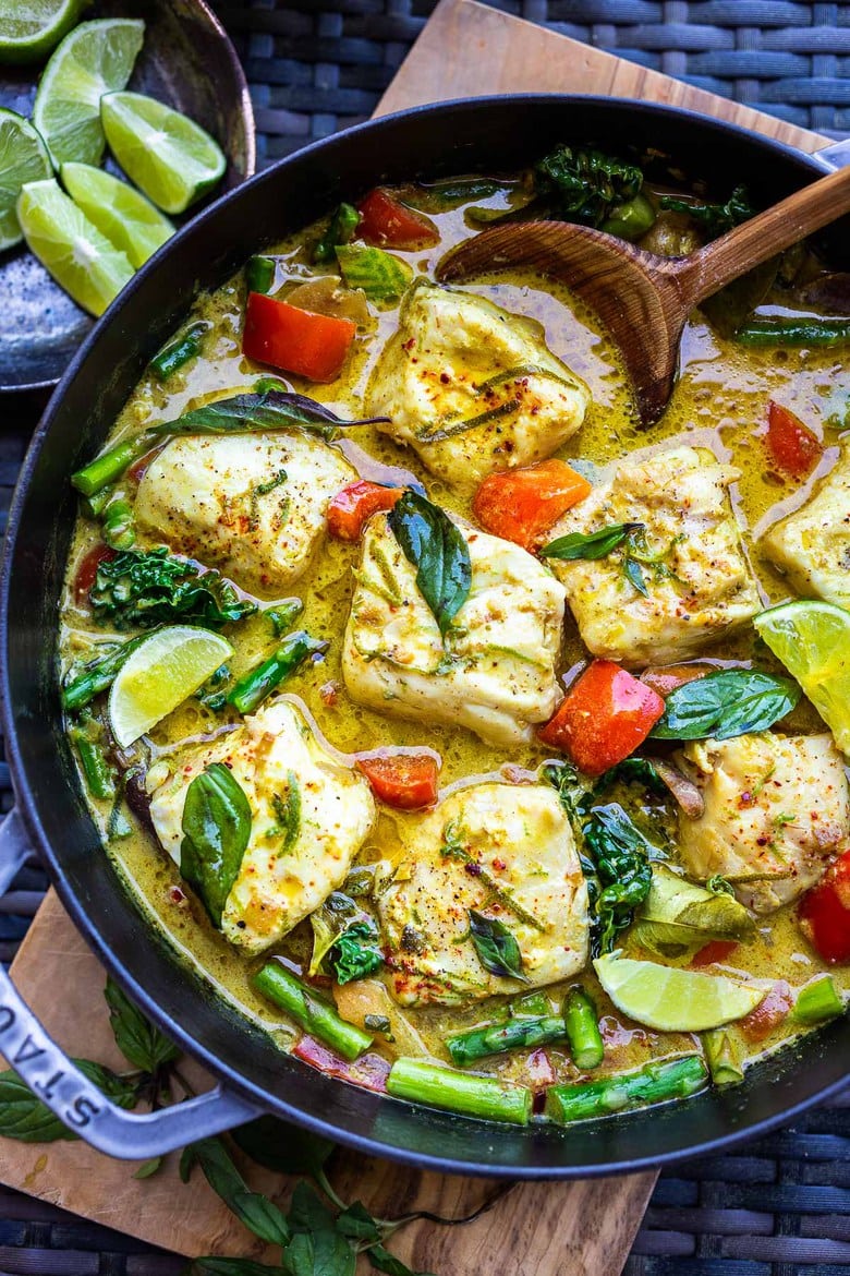 dutch oven with colorful thai fish curry and wooden spoon- fish topped with lime zest, basil, and lime wedges. 